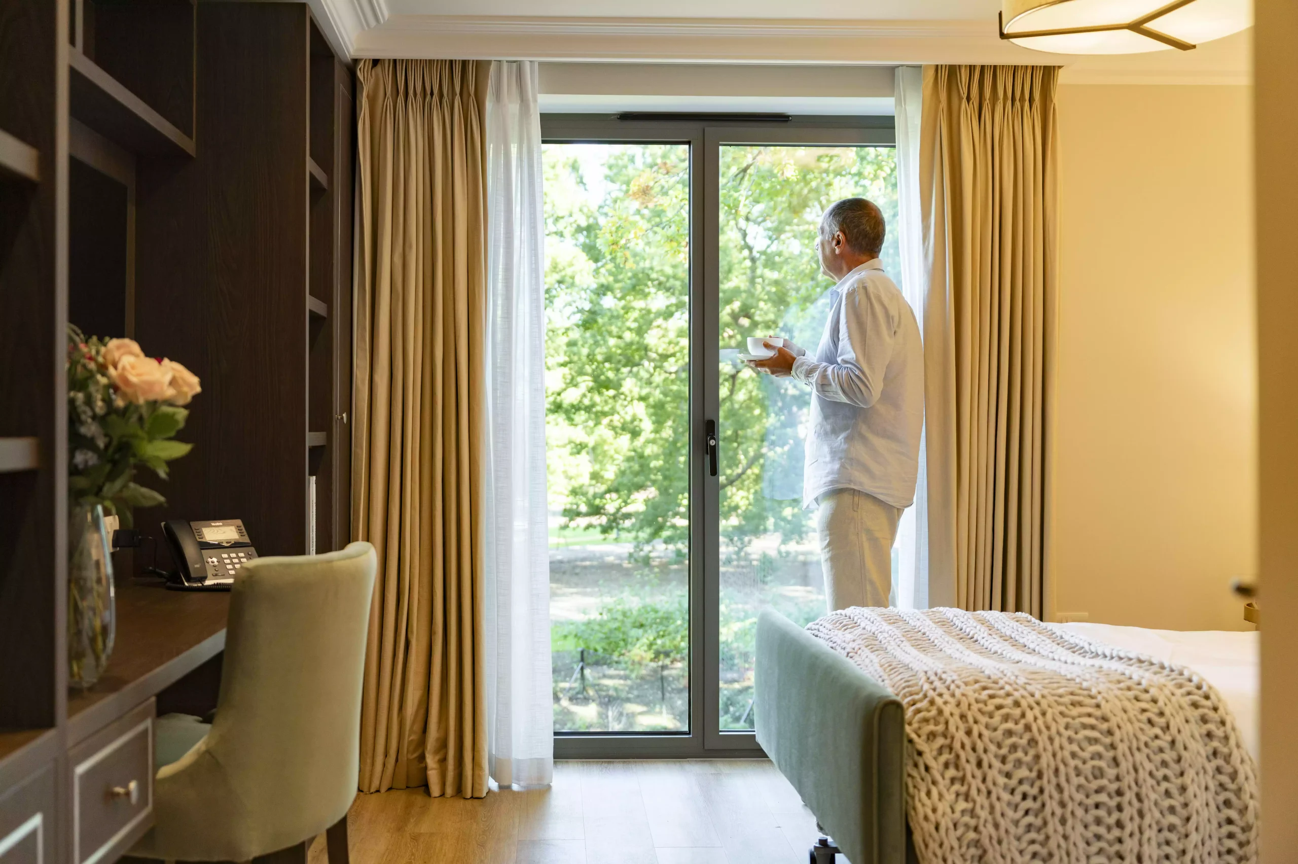 A standing man holding a cup of tea looking outside the window