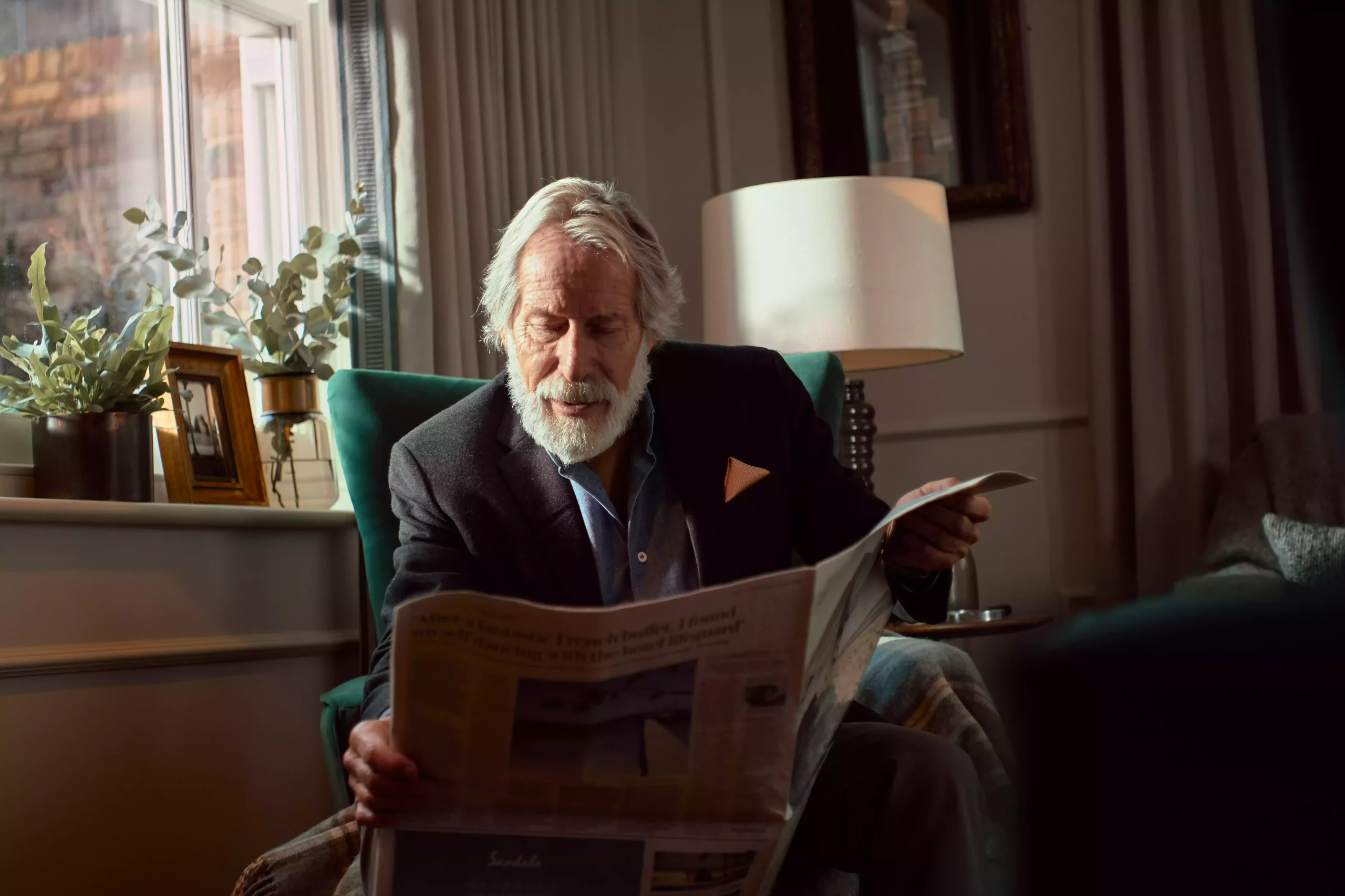 An old man sitting on a chair reading the newspaper.